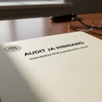 Puidust laual lebab paber kirjaga „AUDIT JA HINNANG koosatakse Ehitusseadustiku alusel”, mille kõrval on prillid.